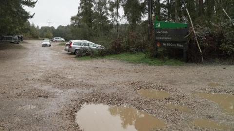 Deerleap car park in the New Forest on a dull day - six cars are parked and there is a number of potholes in the foreground, filled with rainwater