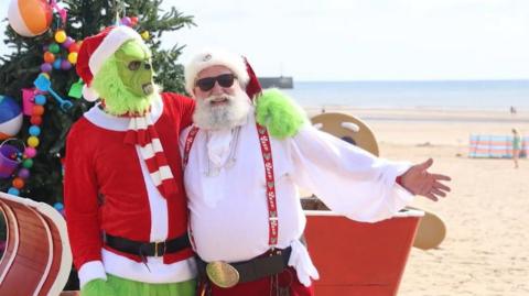 A man dressed as Santa, with a red and white Christmas hat, sunglasses, a white shirt, red braces and a big belt with a gold round buckle, puts his arm round a man dressed as the Grinch, with green fur and a red hat, jumper and scarf. They are standing on a golden beach on a bright summer's day with the blue sea in the background. A Christmas tree and a red sleigh are behind them.