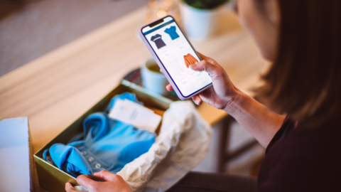 Woman looks at smartphone showing clothing with a package on the table showing a blue top