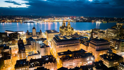 Aerial view of Liverpool city centre showing the three graces and the River Mersey with Wirral in the background. It is taken in the evening with all the building lit up.