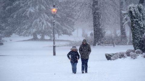 Two people walk through a snow-covered park while thick snow continues to fall