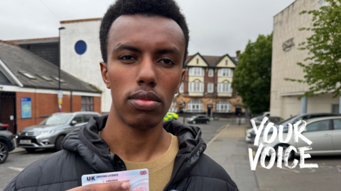 Robert is facing the camera and holding his drivers licence in his hand which he recently passed. He is standing on a residential street in Croydon with parked cars behind him.