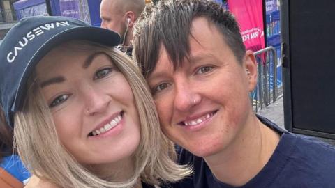 Close up of two womens' faces. The woman on the left has shoulder-length blonde hair, a nose ring and is wearing a navy baseball cap with the word CAUSEWAY embroidered across the front. The woman on the right has short dark hair, a nose ring and a navy t-shirt. Flags, speakers, crowd barriers and a gazebo are behind them suggesting they are at a public event.