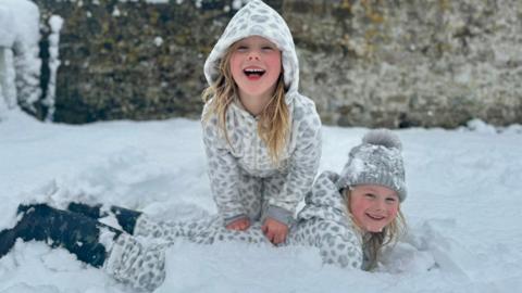 Two young girl twins dressed in grey pyjamas and black wellies laying on the floor in deep snow. 