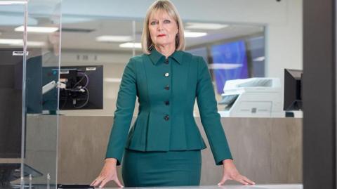 Joy Allen staring into the camera with her hands resting on a surface in front of her. She's got shoulder-length blonde hair and is wearing bright red lipstick. She's wearing a dark jacket that some people would say is blue and others green, with a matching skirt. She's standing in what looks like a police station. There are computers behind.