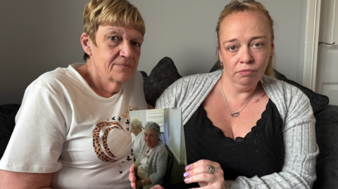Karen Day on the left wears a white shirt with a pattern on it, while sitting next to her daughter Leanne Day, who is wearing a black shirt and a grey cardigan and holding up a photograph of their mother and grandmother, Joyce Sinclair. 