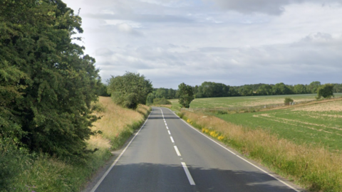 A general view picture of the A361 near Burford. There is no traffic on either side of the road and there is a large tree and hedgerows on the left. There are fields on the right.