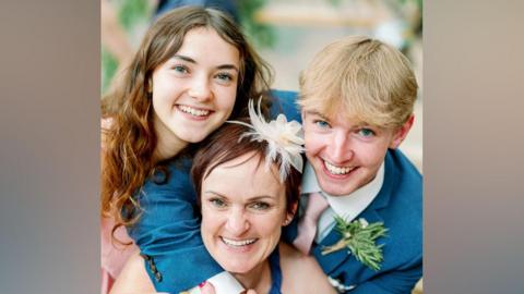 Close-up of three people grouped closely together indoors. In the foreground, one person with short dark hair wears a light-coloured outfit and a decorative white feather hair accessory, along with bracelets and rings. Two younger-looking individuals lean in from either side, both wearing blue suits; one has long brown hair parted in the middle, and the other has short light blond hair and a boutonniere pinned to the suit jacket. All three are positioned tightly together with arms wrapped around each other, against a softly blurred background with greenery."