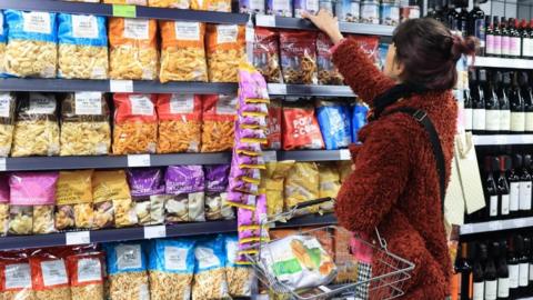A female shopper stands facing shelves with crisps on, holding a basket