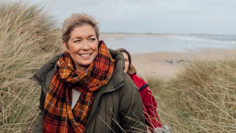 Two women walk through sand dunes on a beach. Both are wearing weather coats and scarves.