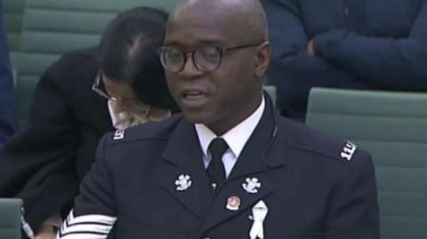 Inspector Tola Munro wearing his police uniform and glasses while sitting on green chairs in a committee meeting.