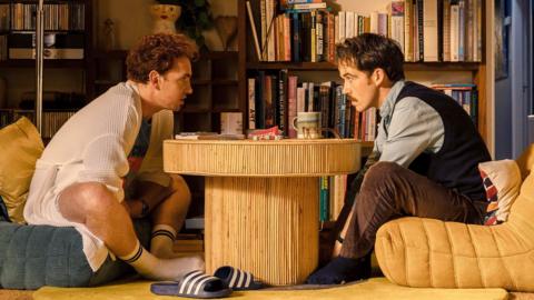 Two men sit on opposite ends of a board game. They look like they're talking