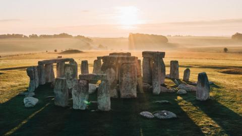 Stonehenge seen with the sun behind and sun beams streaming through the large stones placed in a circle and lighting up the hills and fields behind.