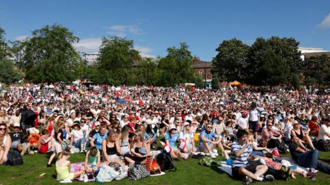 Several thousand people are seated on the grass on Devonshire Green facing centre-left of the camera, looking at a large screen.