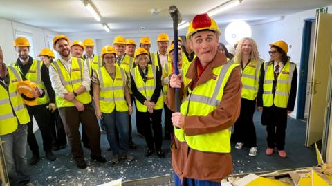 A man wearing light clown's makeup, a yellow builder's helmet and a yellow high visibility vest brandishes a mallet after knocking down a wall in a community centre, a quirky expression on his face. A group of around 20 people are standing behind him, smiling, also wearing construction gear.