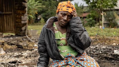 A woman in an orange head scarf looks at the camera holding her head with one hand.