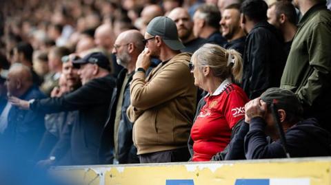 Manchester United fans look on during the Premier League 2026/27 match between Hull City and Manchester United at MKM Stadium. One fan in the foreground appears to have their head in their hands. 