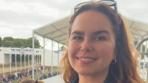 Molly is pictured outside at an event. She has long brown hair and is smiling at the camera. She is has small hoop earrings in and sunglasses on her head.