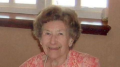 A brown-haired woman smiling. She is wearing a pink outfit with a pearl necklace and pearl earrings