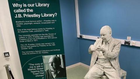 A sign on the left explains why the University of Bradford's library is named for the writer J B Priestley and next to the sign is a white statue of the author seated and holding a pipe in his hands