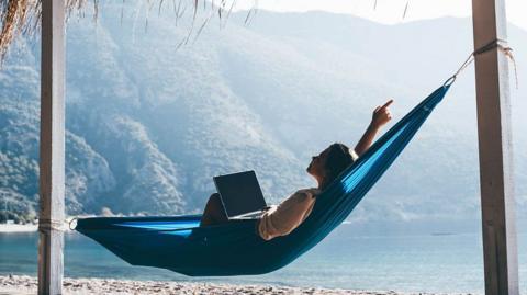 A person lying in a hammock with their hand in the air and a laptop on their lap.