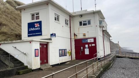 Sheringham Lifeboat Station is a square-form block building featuring windows and red doors. It has aerials on the roof, and a large sign bearing the RNLI flag, and the word Lifeboats. It is on the promenade, with railings on the edge, towards the shingle beach. Behind the station is a sloping cliff.