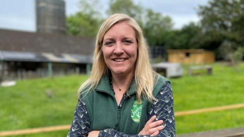 Milly Fyfe stands on her farm in front of a wooden fence. Agricultural buildings and a field are in the background. She has blonde hair and is smiling into the camera. She is wearing a green fleece zip-up vest over a black patterned blouse.