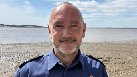 A man wearing a blue shirt with epilates. He has short hair and a beard and is standing on a shoreline.