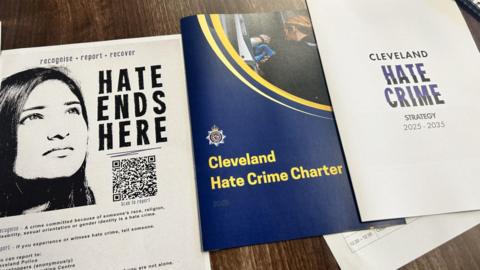 Three A4 leaflets on a dark wooden table. The first black and white leaflet has the face of a woman with the words "hate ends here" with a QR code below. The next leaflet is dark blue with the words "Cleveland hate charter" in yellow. The third white leaflet has the words "Cleveland Hate Crime Strategy 2025-2035" on the front.