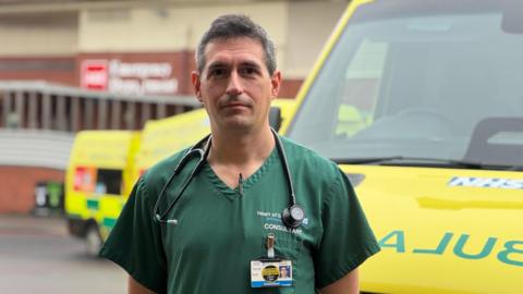 A man with dark grey hair stands in green hospital scrubs by an ambulance, in front of a hospital building.