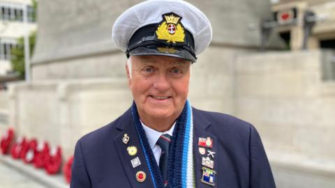 Keith is wearing a navy blazer adorned with military medals and badges, a white naval-style cap and a blue scarf. He stands smiling in front of the Hull Cenotaph - a large stone war memorial. Red remembrance wreaths are laid at the base of the memorial behind him, and buildings and trees are visible in the background.