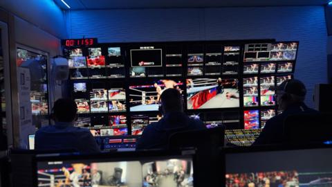 Backstage at WWE. There are many camera screens each showing a different angle of the match. Three men watch.