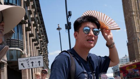 A man in sunglasses holds a fan to shield himself from the UK sun during this week's hot spell