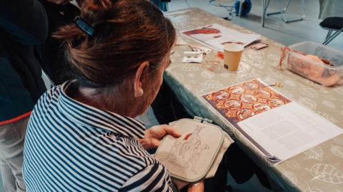 A photo of a woman embroidering. She has her brown hair tied up in a clip and has her back to the camera. She is wearing a black and white striped top. The thread on the embroidery is red. In front of her is a table with papers on.