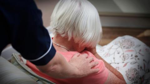 A white haired woman with her back to the camera is comforted by a hand on her shoulder from a carer