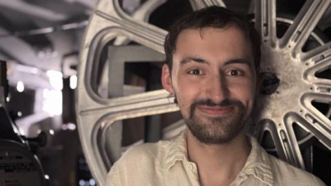 Patrick smiles at the camera, in front of a large wheel that holds film in the projection room. He has dark hair and a short dark beard, and wears a white loose shirt. The room behind him is dark and full of machinery