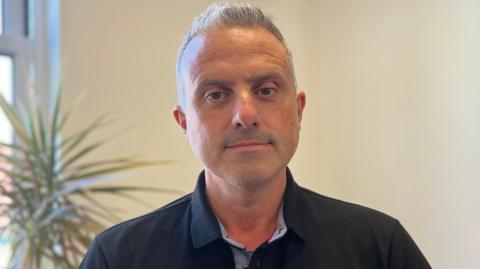 A man wearing a black collared shirt with a plant and plain wall behind.