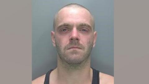 Arran Brown in a police mug shot. He has shaved head and stubble and is wearing a black vest.