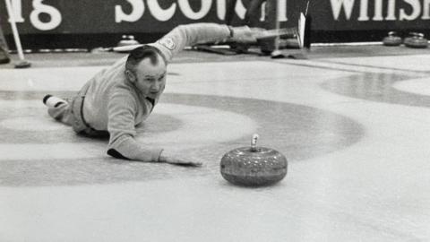 Hammy McMillan lying on his stomach on the ice with his right arm resting on the ice, having just thrown the curling stone- his left arm is held in the air