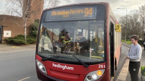 A red single-decker bus at a bus stop. Its destination board reads: "South Woodham Ferrers Asda 94". A passenger waits to board.