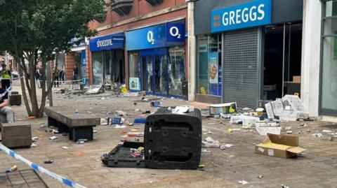 Rubbish, bins and rubble strewn across the ground in front of damaged shops, including shoezone, O2 and Greggs, some with their windows smashed.