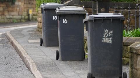 Three black wheelie bins have been left out for collection in front of low stone wall on a kerbside. 