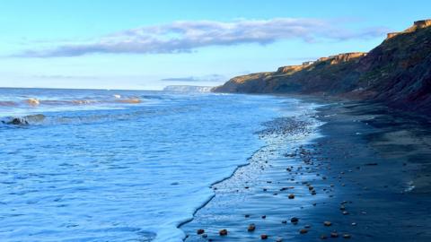 An empty sandy beach with large pebbles on the shore line. There is a low cliff behind the beach with waves breaking on the sand. In the distance you can see the white cliffs of a headland and the sky overhead is blue with a large white cloud.