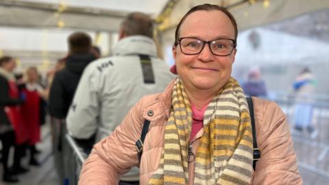 Anna Gillham smiles at the camera in a tent where soup is being served. She is wearing a pink coat with a pink jumper and a striped scarf. She has glasses and her brown hair is tied into a ponytail.