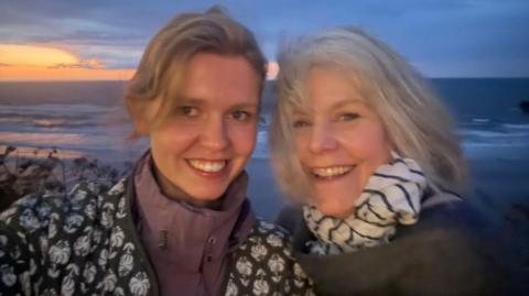 Two smiling women. The woman on the left has blonde hair and is wearing a pink zip up jacket with a brown and white jacket on top. The older woman on the right, has shoulder length blonde hair and is wearing a brown jacket with a white and brown striped scarf.