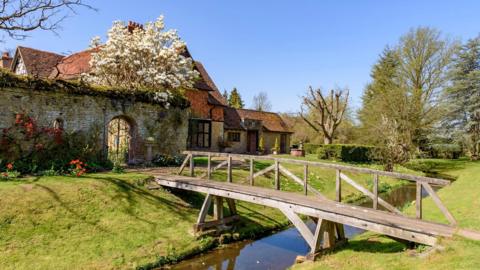 Great Tangley Manor, which is an old, stone house with a pointed, red-tile roof. The property is surrounded by manicured grass and other plants, whole a narrow wooden bridge crosses a moat around the perimeter.