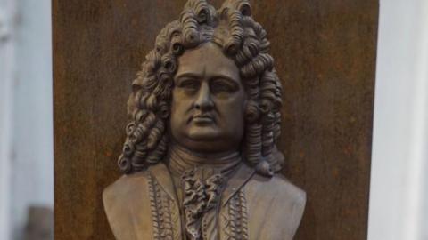 A bronze bust which will be placed on a plinth. The man depicted has long curly hair, and is wearing a ruff and ornate jacket. 