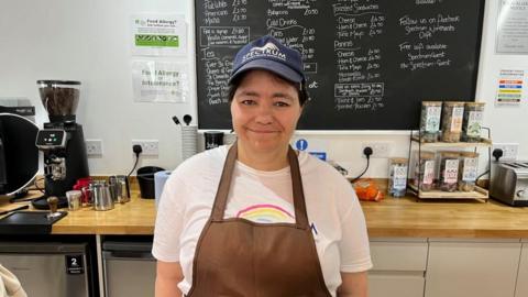 A woman in a T-shirt, cap and an apron standing in front of a coffee machine and counter