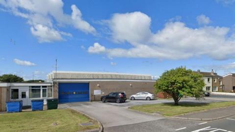 A Google Street View screenshot of the entrance to Distington Ambulance Station: a low, concrete building, with a large garage-style entrance painted in blue. In the foreground, to the right, is a tree standing on a lawn. To the left, another area of green, with industrial bins in front of a white door and window.