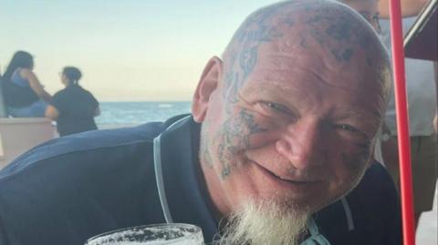 A close-up view of Stephen seated at what appears to be a seaside restaurant, café, or bar. He is leaning forward toward the camera and holding a glass of beer in one hand.
He has tattoos visible on the head, face, fingers, and hand. A long white goatee beard extends from the chin. He is wearing a dark collared shirt with light-coloured trim around the collar and shoulders. Partially visible white text on the shirt.